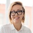 Happy middle aged businesswoman in eyeglasses sitting by her workplace in office and looking at camera with toothy smile
